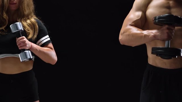Athletic man and woman flexes their hands with dumbbells, training their biceps on a black background in studio. They stand at different edges of the frame. Center for text