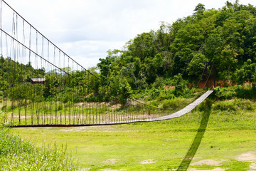 Rope bridge with wood planks across over the river and mountain forest in Thailand