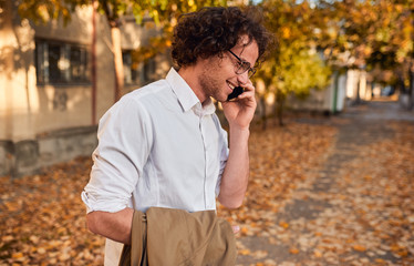 Side view shot of handsome businessman posing on the autumn street while walking outdoors and using smartphone for calling. Smiling man with curly hair wearing spectacles talking at his mobile phone.