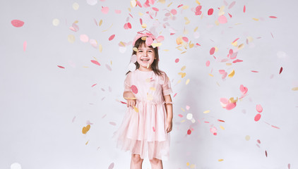 Obraz na płótnie Canvas Happy cute little girl wearing pink dress in tulle with princess crown on head isolated on white background playing with confetti. Joyful pretty little girl celebrating her birthday party, having fun