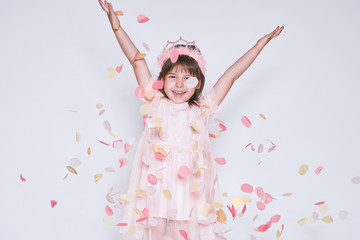 Obraz na płótnie Canvas Funny little girl wearing pink dress in tulle with princess crown on head isolated on white background rise hands up enjoy confetti. Pretty happy little girl celebrating her birthday party, having fun