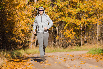 Obraz premium Young adult man running on rural road in sunny day. Sportsman in tracksuit with hood and sunglasses. Outdoor workout in golden autumn time. Daily active lifestyle. Enjoying sport. Front view.