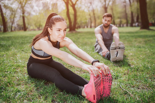 Attractive girl is sitting on grass in park and stratching. She is looking straight and smiling. Her boyfriend isstretching as well and looking at her partner.
