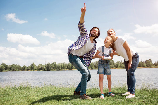 Nice picture of young family looking up in the sky. Guy is pointing up. Girl has a control panel on hands. Parents are holding their daughter's back. They are happy.