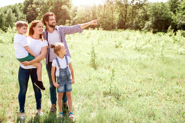 Fototapeta premium Nice picture of family standing together on meadow. Mom is holding her son on hands. Girl is standing besides her parents. Guy is pointing down further.