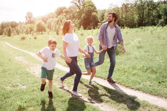 A picture of full family walking and jumping together on the road. THey are going through green meadow. They are laughing.