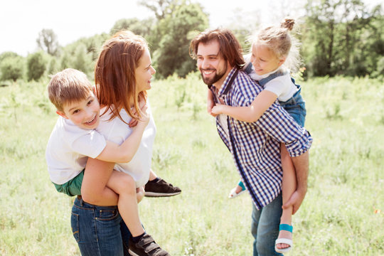 Funny moments of good family. They are standing outside on meadow. Parents are looking a each other and smiling. They are holding their kids on their back. Children are having some fun as well.