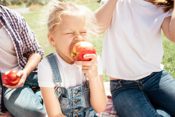 A picture of small girl biting an apple. She is doing that with pleasure. Her parents are sitting behind her. Guy has an apple in hands.