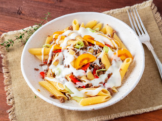 Penne pasta with yellow tomatoes, vegetables, mincemeat, white sauce on dark wooden background, side view, close up