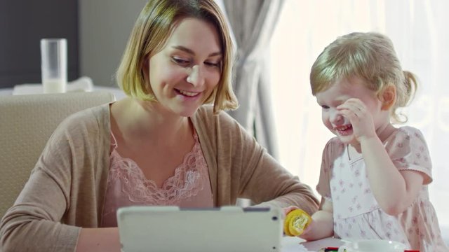 PAN of happy young mother and cute toddler girl sitting at table and laughing while cartoons on tablet