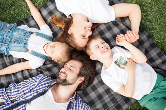 Gorgeous members of family are lying on the blanket on the grass. Man is looking at daughter. Woman is looking at son. They are smiling and laughing. Family is speding time together.