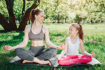 Girls are sitting on carimate in lotus pose and loking at each other. They are smiling. Girls are excercissin outside in park in a beautiful sunny day. Yoga and Pilates Concept.