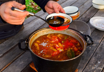 Cooking food in nature. Men, the cook pours a scoop of soup from the pot into a crane bowl. Tomatoes, pepper, diet. Homemade delicious lunch