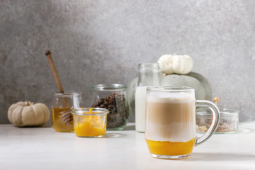 Glass of pumpkin layered spice latte with pumpkin puree, milk foam and cinnamon standing with ingredients in jars and decorative white pumpkins on white marble table with grey wall at background.