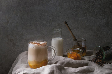 Glass of pumpkin layered spice latte with pumpkin puree, milk foam and cinnamon standing with ingredients in jars and decorative white pumpkins on crumpled table cloth with grey wall at background.