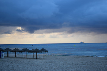 Sandy beach of the Mediterranean coast of Spain with parasols, palm trees and a view of a sailing ship away in cloudy rainy weather