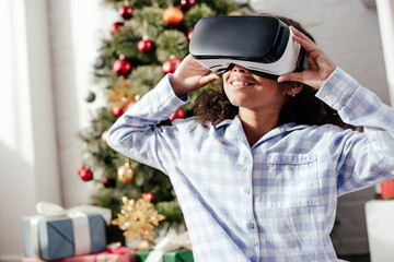 cheerful african american child in pajamas using virtual reality headset at home, christmas concept