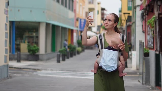 Mom walks on the street with her baby in a sling while traveling to Spain and takes pictures of the sights on her mobile phone