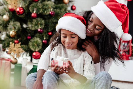 happy african american woman and daughter with gift in santa claus hats at home, newy year and christmas concept