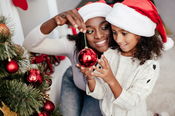 Smiling African American woman in Santa Claus hat helping daughter to decorate Christmas tree at...
