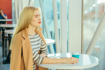 Young elegant positive blond business woman in beige coat with red lips drinking coffee, reading book, sitting at round table indoor by break time in modern office center.