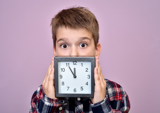 Surprised Young Boy Holding And Showing A Clock And Looking At The Camera. Clock Showing Nearly 12.