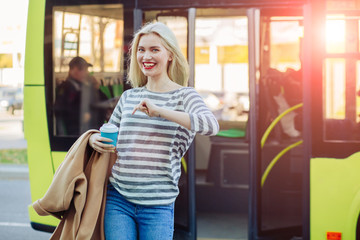 Young positive blond hipster woman passenger with coffee cup getting off the yellow bus in...