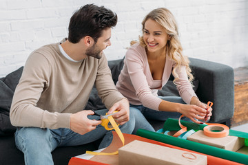 smiling couple preparing christmas gift box together, holding ribbons and looking at each other at home