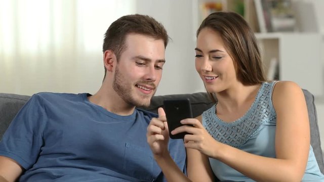 Happy couple talking about smart phone content sitting on a couch in the living room at home