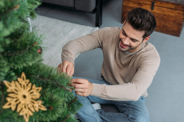 high angle view of smiling handsome man decorating christmas tree with baubles and sitting on floor at home