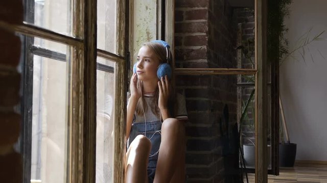 Portrait of young attractive girl on window background is listening to music with headphones. A beautiful teen girl sits near a window with headphones listening to music from smartphone