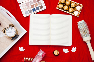 Personal things girls on a red background. An open notebook in the center, near an alarm clock, a palette of eye shadows, souvenirs, a bottle of perfume, red gloves and sweets