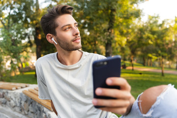 Fototapeta premium Young guy sitting in park outdoors using mobile phone listening music with earphones.