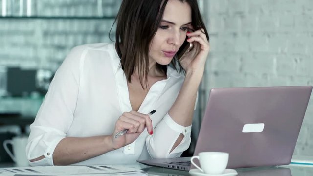 Pretty businesswoman talking on cellphone and comparing data on laptop and documents in the office
