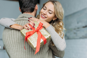 beautiful young woman with gift box embracing her boyfriend at home