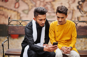 Two indian stylish mans friends in traditional clothes posed outdoor, sitting on bench and looking at phone.