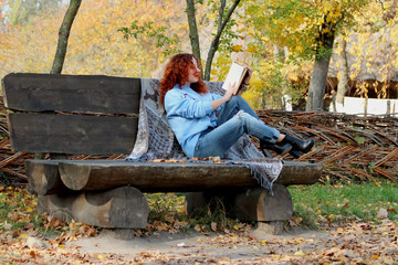  Beautiful Woman with red hair in the autumn park. sits on a bench and reads a book. Autumn background. Nearby is a warm blanket with deer