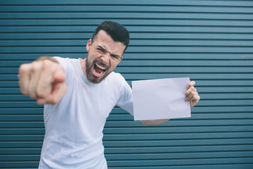 Guy is standing and posing. He is looking and pointing on camera. Also man is holding blank piece...