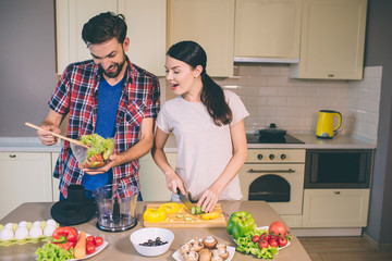 Excited girl stands at table and cuts yellow pepper. She looks at bowl with salad the guy is holding. He mixes ingredients with wooden spoon. They cook together.