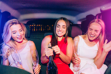 Beautiful girls are sitting in car. Brunette is opening bottle with alcohol. Blonde girls are very...