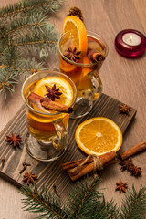 Two glasses with delicious fragrant tea of mulled wine with orange and spices on a wooden board with branches of a Christmas tree with a candle and tinsel.