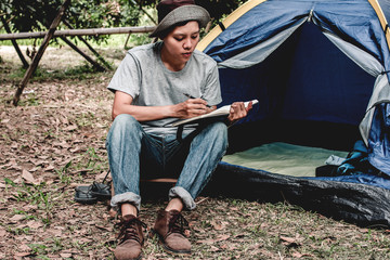 Asian young man sitting is holding a pen writing note of  letter memorize memories on book  in outside the tent. Loneliness camping in forest