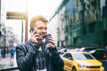 Attractive stylish young man in a leather jacket with a smartphone and take-out coffee in his hands against the backdrop of a large modern city