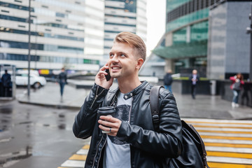 Attractive stylish young man in a leather jacket with a smartphone and take-out coffee in his hands against the backdrop of a large modern city