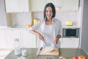 Positive and nice girl stands in kitchen at table and blending together milk with eggs. She likes it. Girl is looking down and smiling.