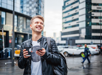 Attractive stylish young man in a leather jacket with a smartphone and take-out coffee in his hands against the backdrop of a large modern city