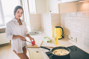 Delightful girl is cutting tomato into pieces. She is cooking breakfast. There are eggs with mushroms frying in pan on stove. She looks on camera and smiles.