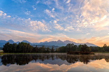 Sunrise shot of peaceful scene of beautiful autumn mountain landscape with lake, colorful trees and high peaks and golden sky in High Tatras, Slovakia.