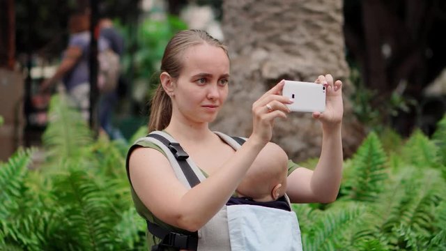 Close up mother mom with baby in a sling travels around the island and takes pictures on a smart phone for social networks