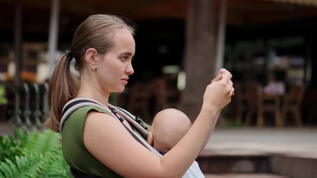 Mom with baby in a sling holding a smartphone in her hands and taking a selfie with the baby